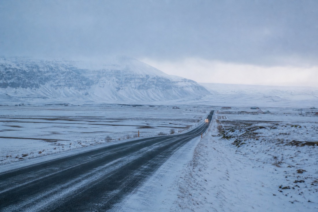 Snow-covered Route 1 running east through south Iceland farmland under flat grey light