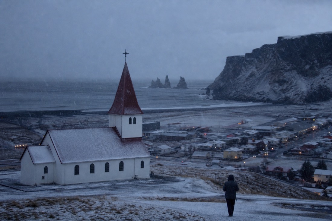 Vik church on its hill above the town in falling snow, black shoreline beyond