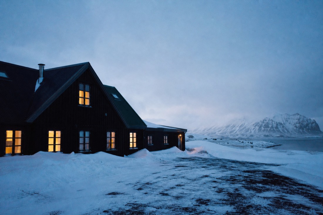 Lit windows of an isolated Icelandic farm guesthouse on Route 1 at dusk with snow drifted against the walls
